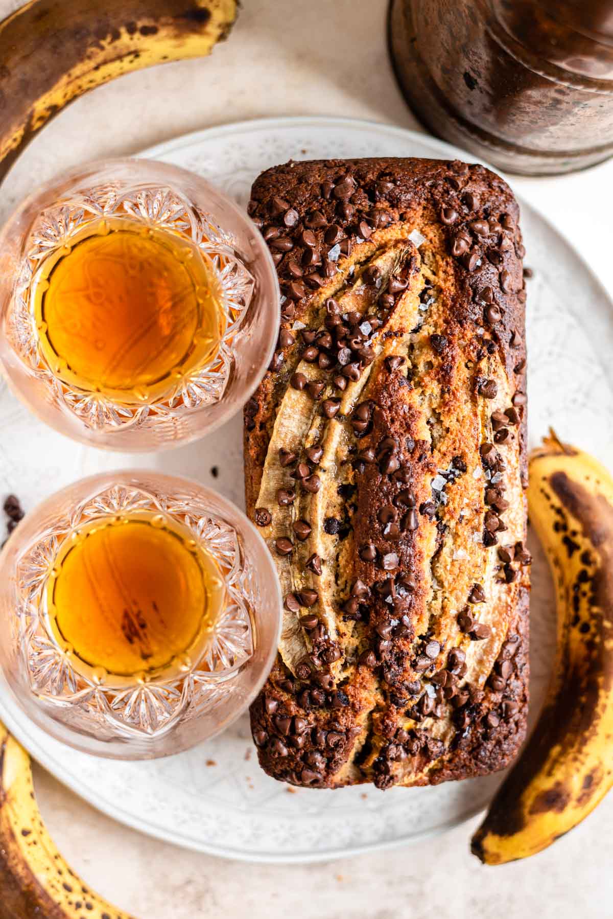 Banana bread on a white plate with two glasses of bourbon on the plate.