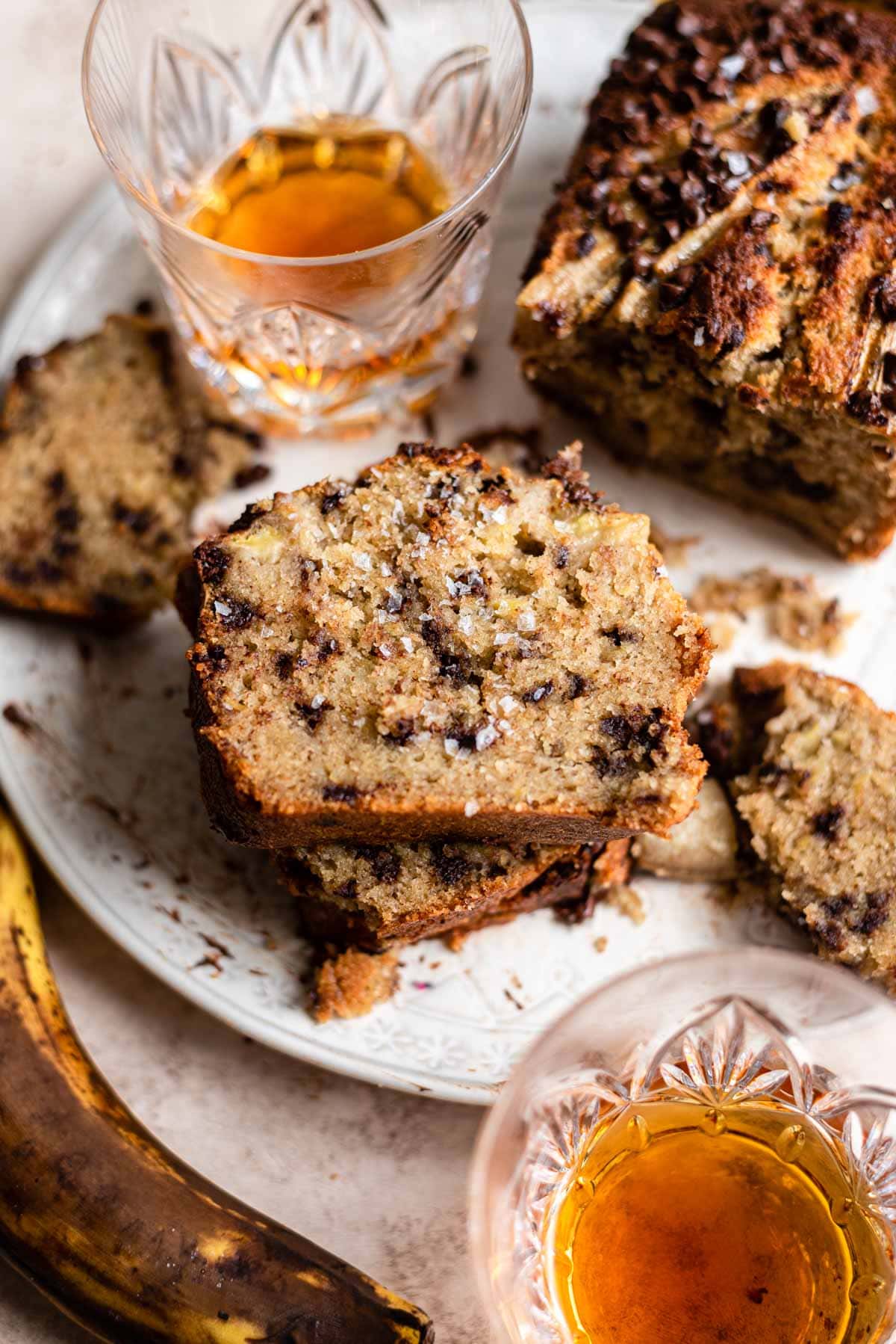 Three slices of bourbon chocolate banana bread stacked on top of each other with two glasses of bourbon above and below the slices on a white a plate.