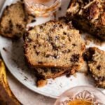 Three slices of bourbon chocolate banana bread stacked on top of each other with two glasses of bourbon above and below the slices on a white a plate.