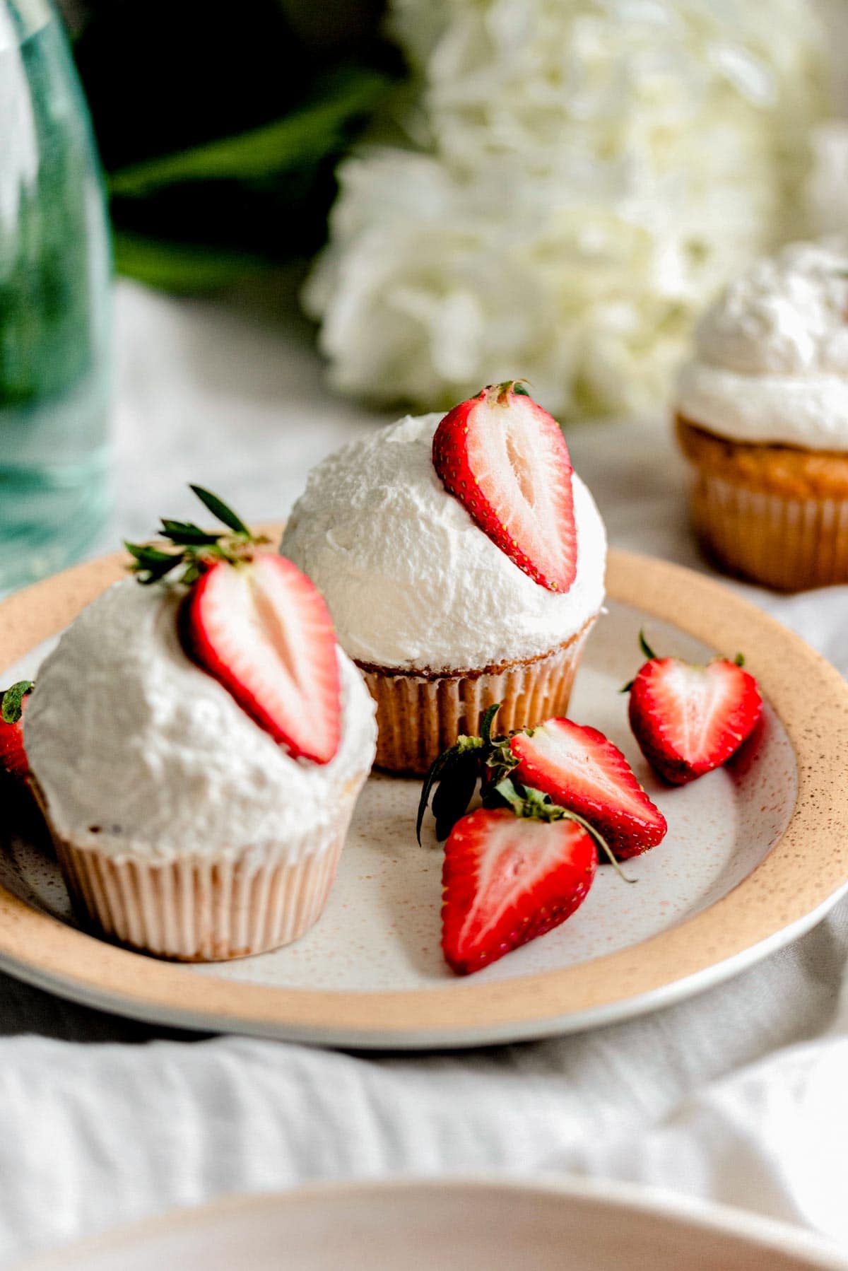 Two cupcakes on a plate with halved strawberries on a white linen table cloth.