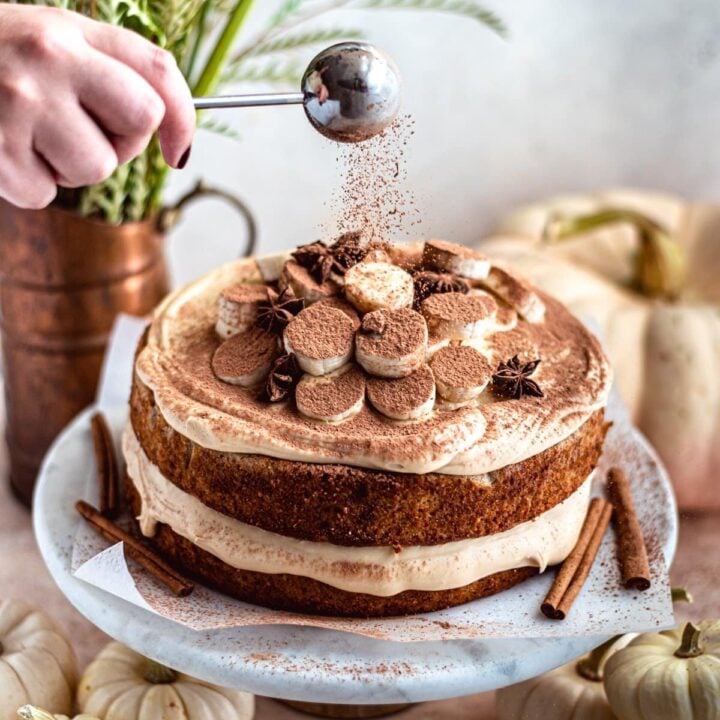 banana layer cake being dusted with chocolate on a marble cake stand.