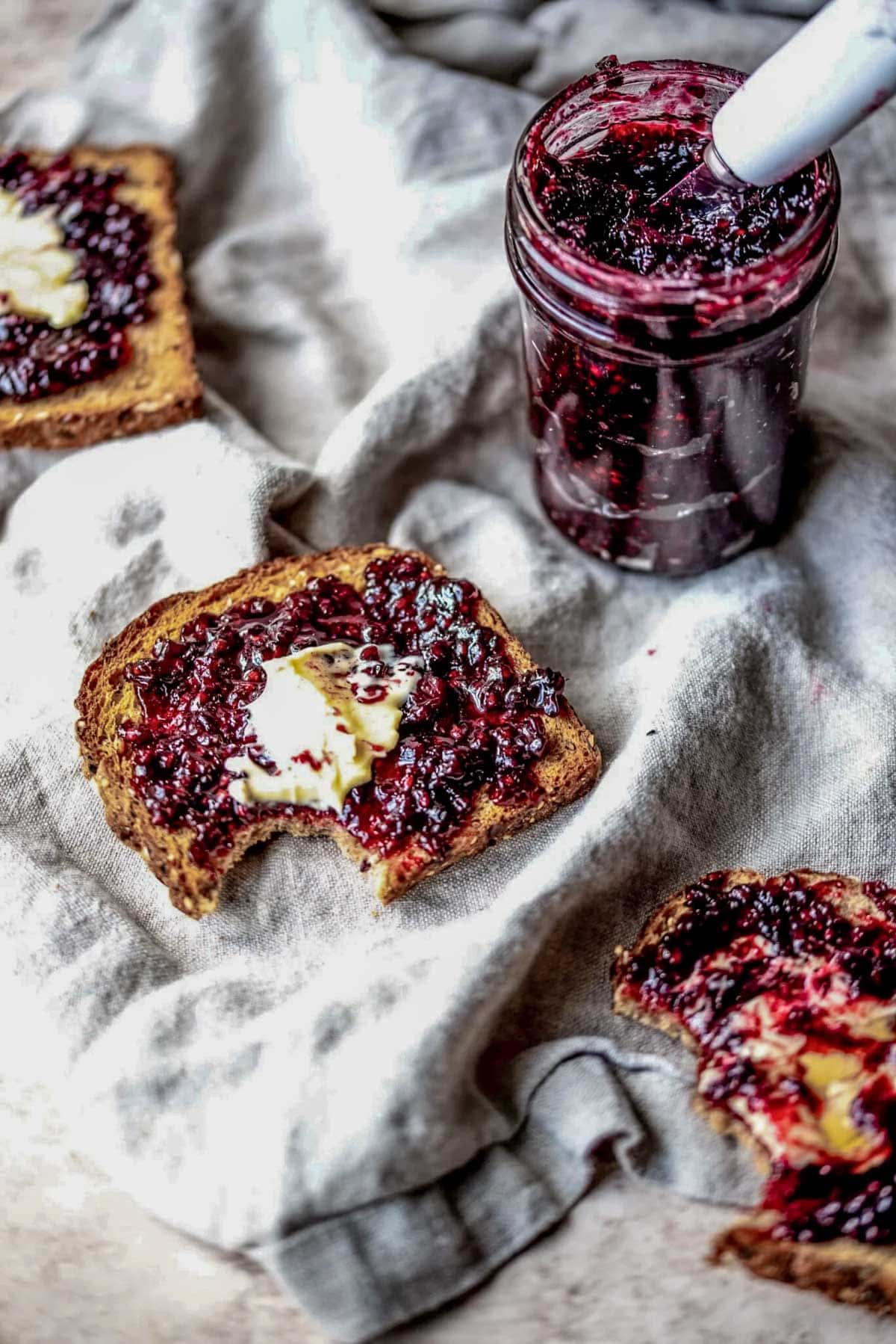 A piece of toast with blackberry jam and butter on top with a bite taken out on grey linen.