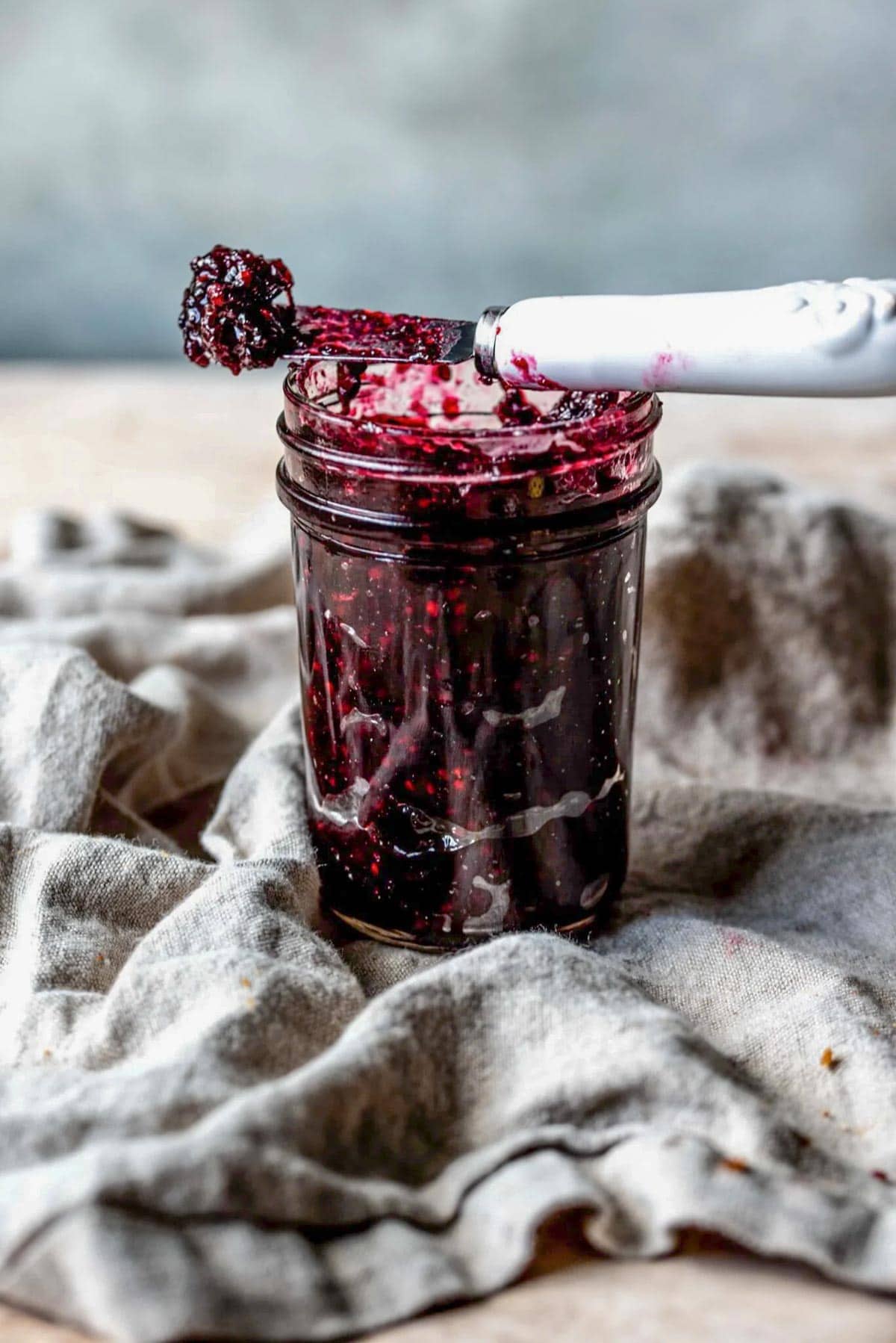 Blackberry jam in small glass jar with a small knife balancing on top of the jar, on top of a grey linen.