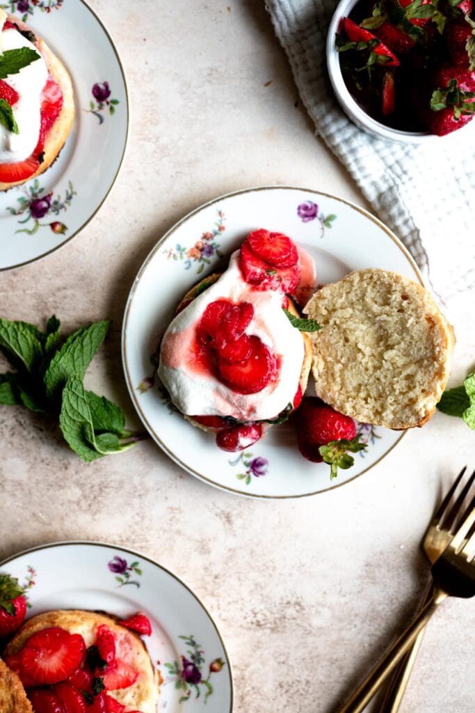 Strawberry shortcake open on a plate with strawberries and mint off to the side and on a brown table.