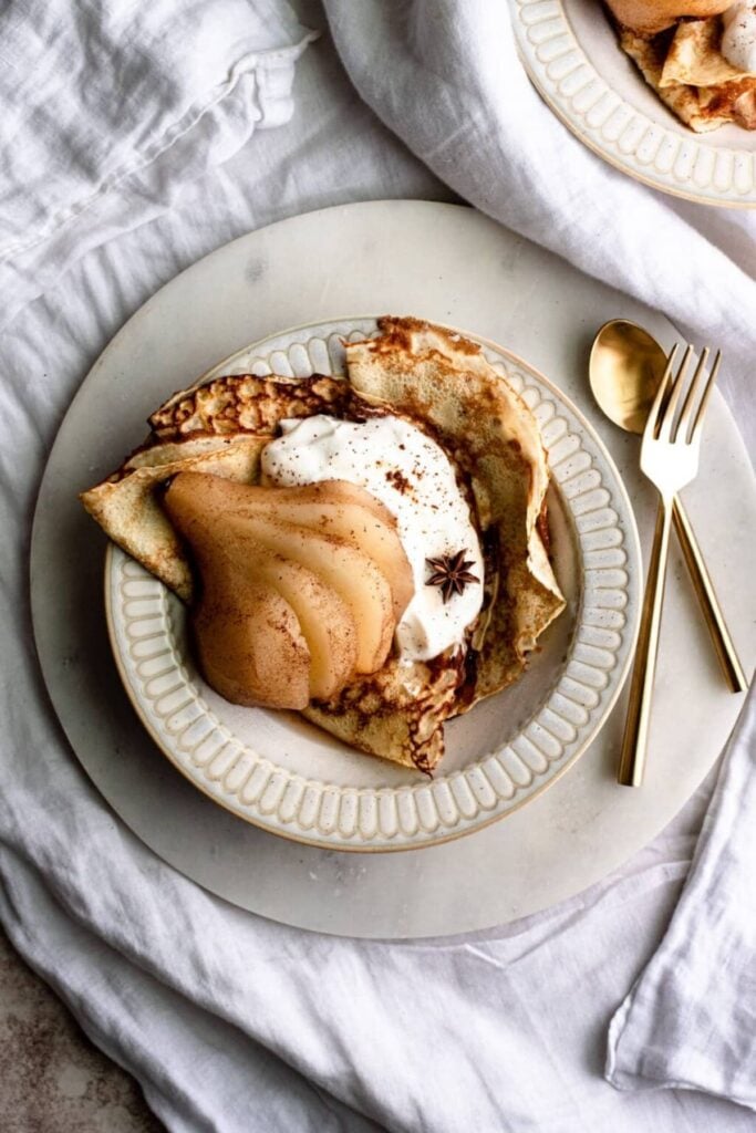 Bourbon Poached pears with folded crepes and whipped cream in a bowl topped with star anise. Fork and spoon are off to the side on a marble platter.