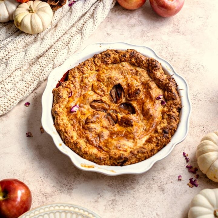 Cheddar apple pie surrounded by apples and pumpkins on a brown table.