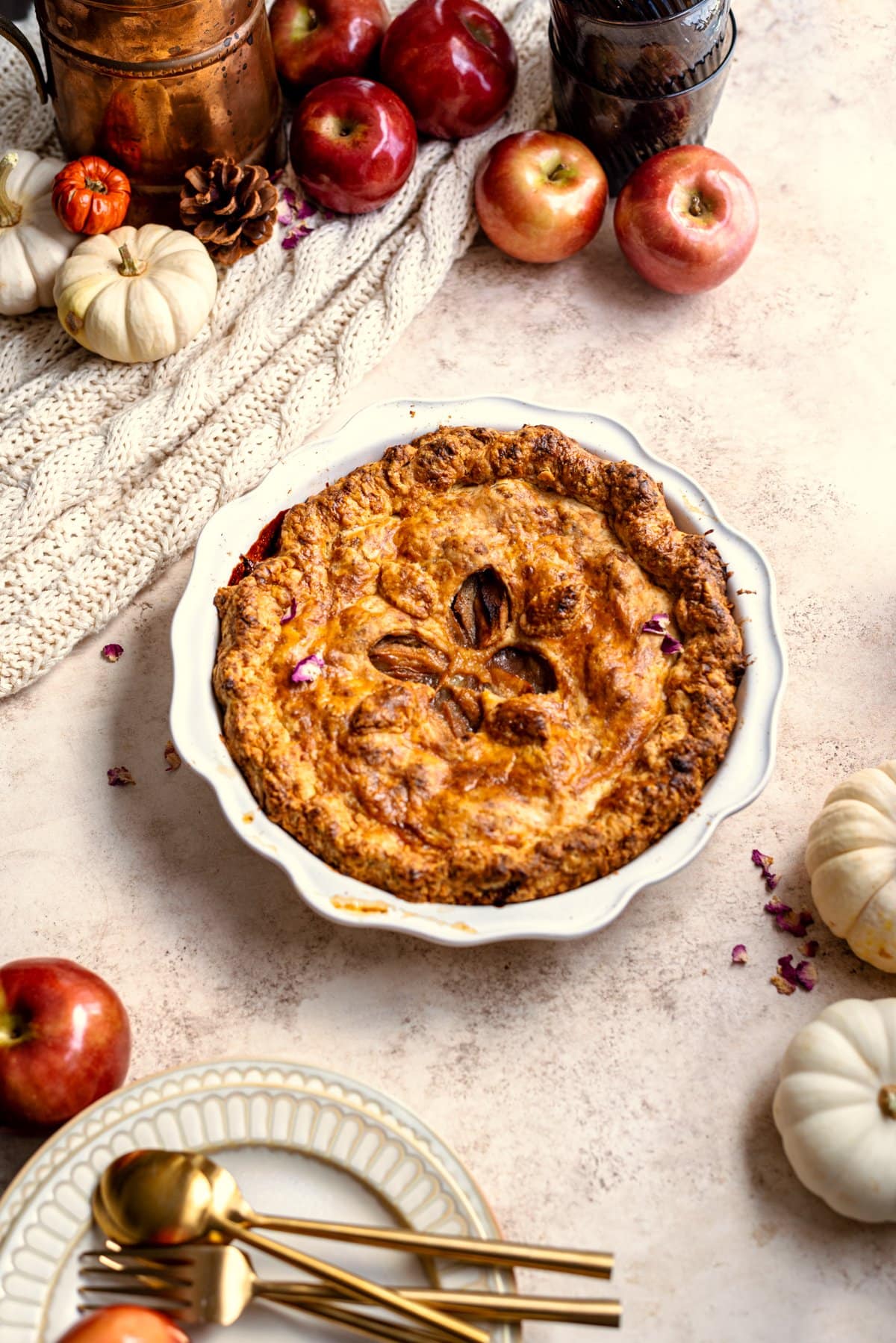 Cheddar apple pie surrounded by apples and pumpkins on a brown table.