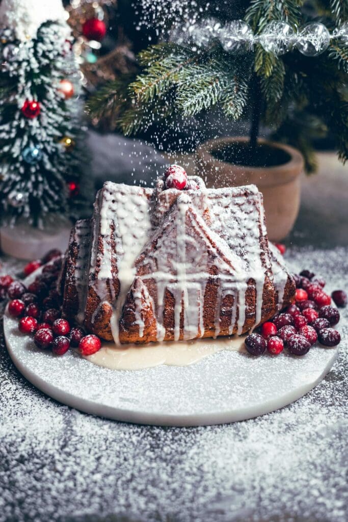 House shipped gingerbread bunt cake on a marble platter with cranberries and being dusted powdered sugar
