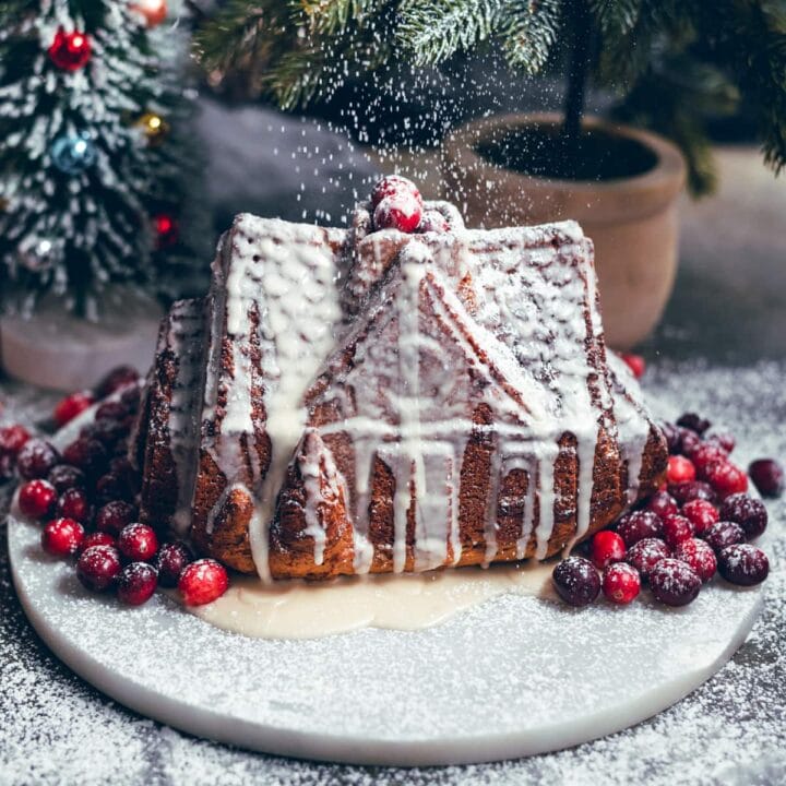 House shipped gingerbread bunt cake on a marble platter with cranberries and being dusted powdered sugar