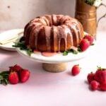 Strawberry pound bunt cake on a marble cake stand with pink strawberry glaze and surrounded by strawberries.