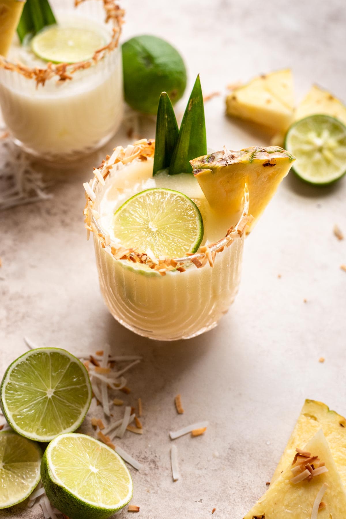 Pineapple coconut margarita garnished with pineapple, lime, and pineapple leafs on a brown table.