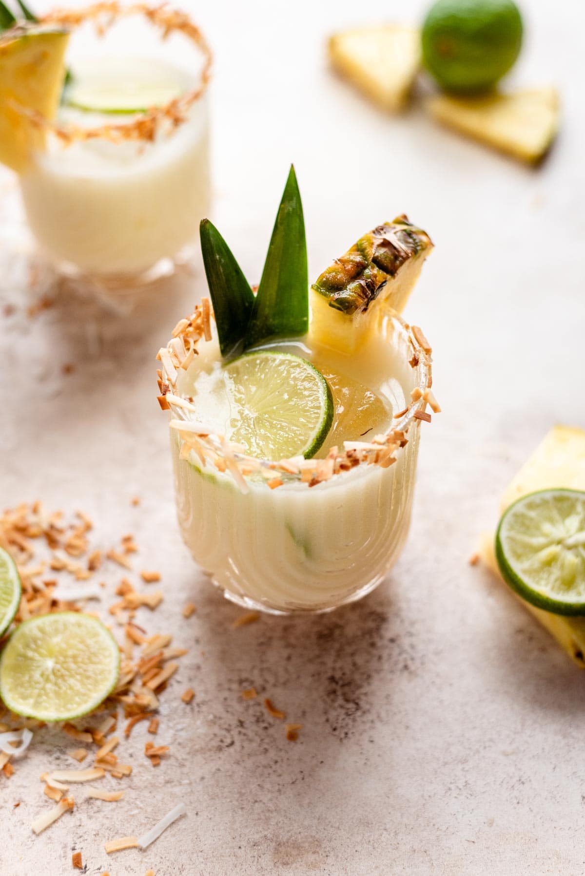 Pineapple coconut margarita garnished with pineapple, lime, and pineapple leafs on a brown table with shaved coconut.