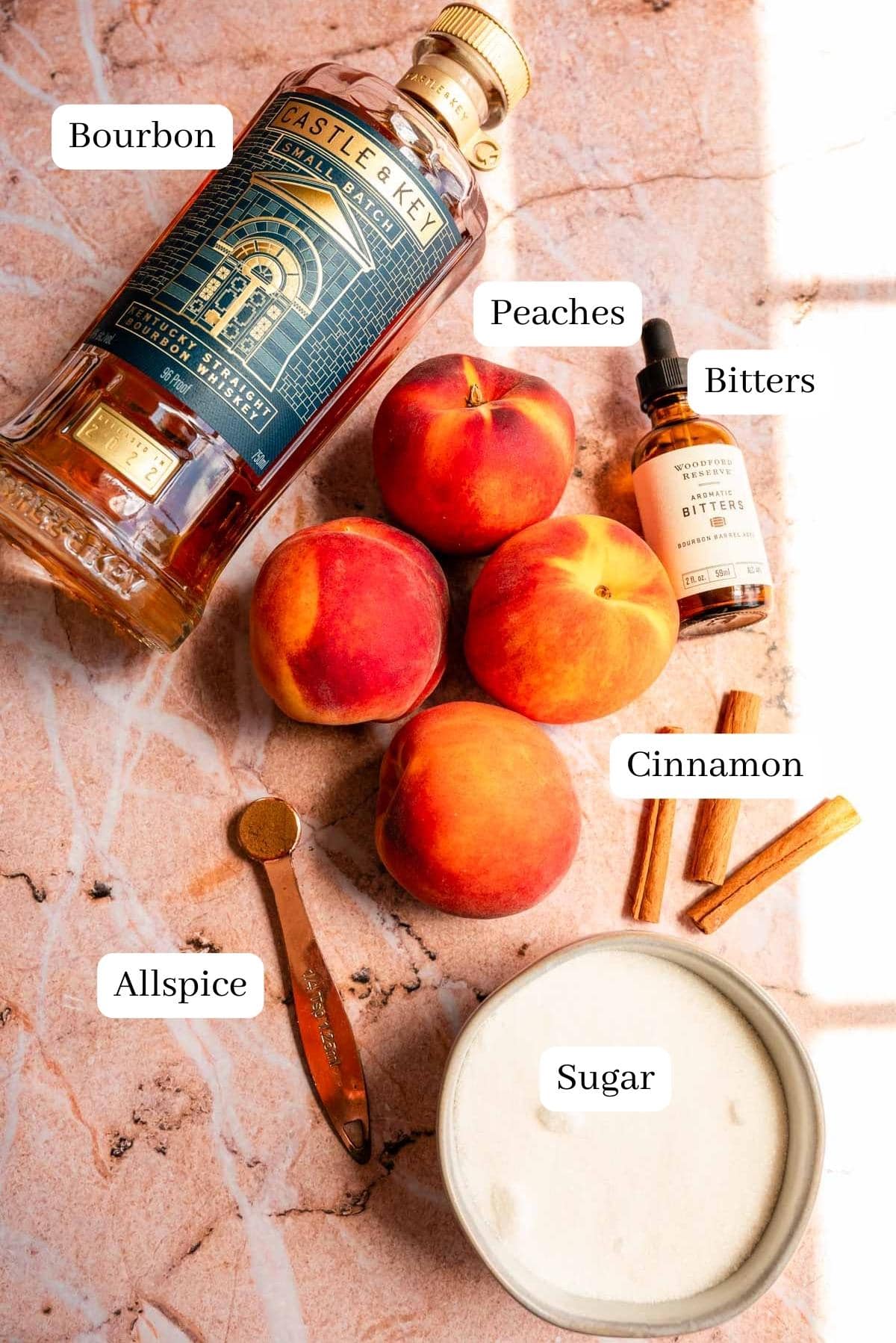 The ingredients, peaches, bourbon, cinnamon, sugar, and bitters laid out on a pink marble table.