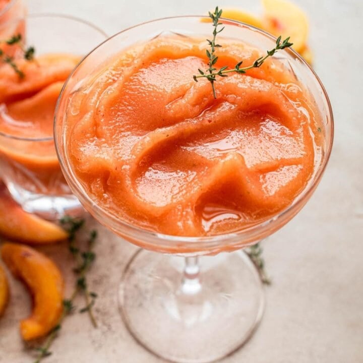 Close up of Bourbon peach slush in a coup glass garnished with fresh thyme on a brown table.