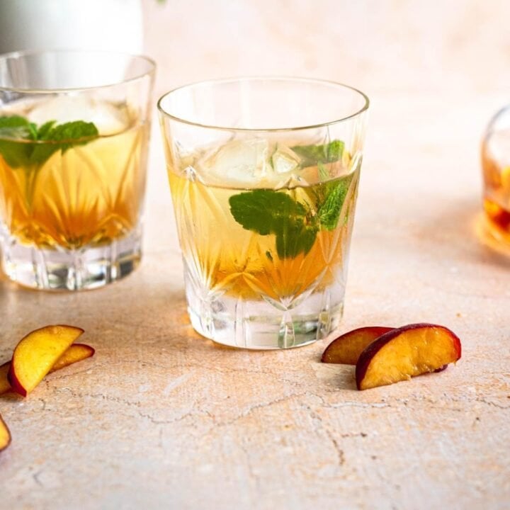 Two glasses of peach old fashioned's on a brown table with peaches and mint.
