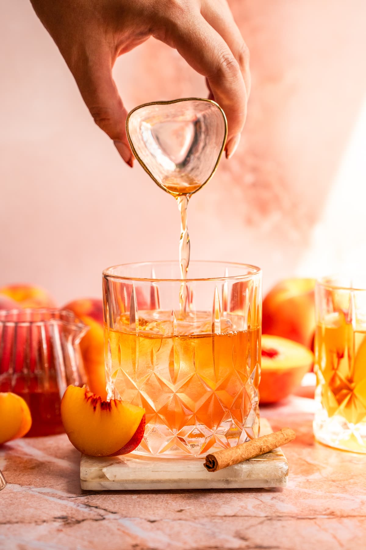 The bourbon being poured into the old fashioned glass with sliced peaches off to the side.