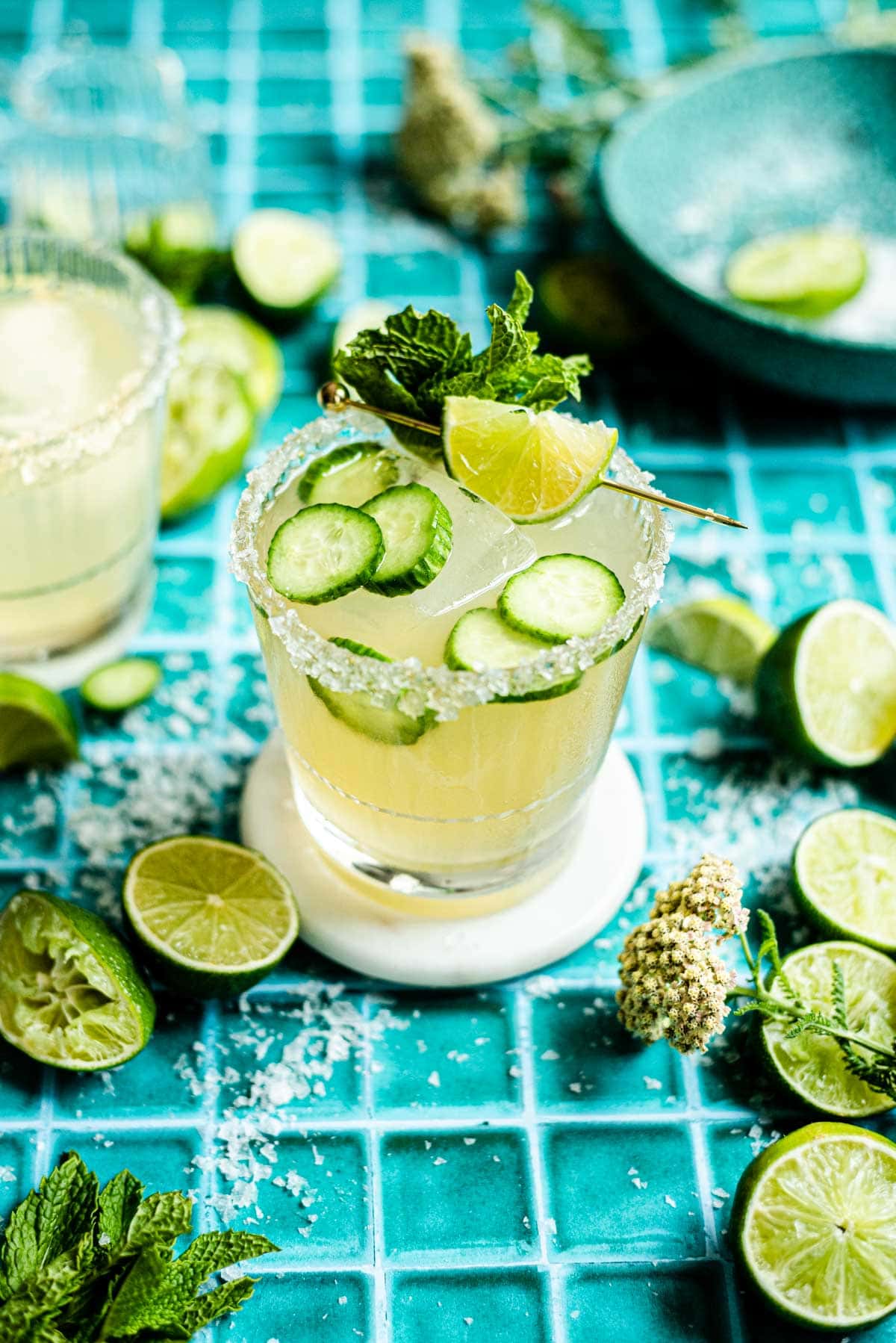 Two Elderflower margaritas on a green tile table surrounded by limes and ice.