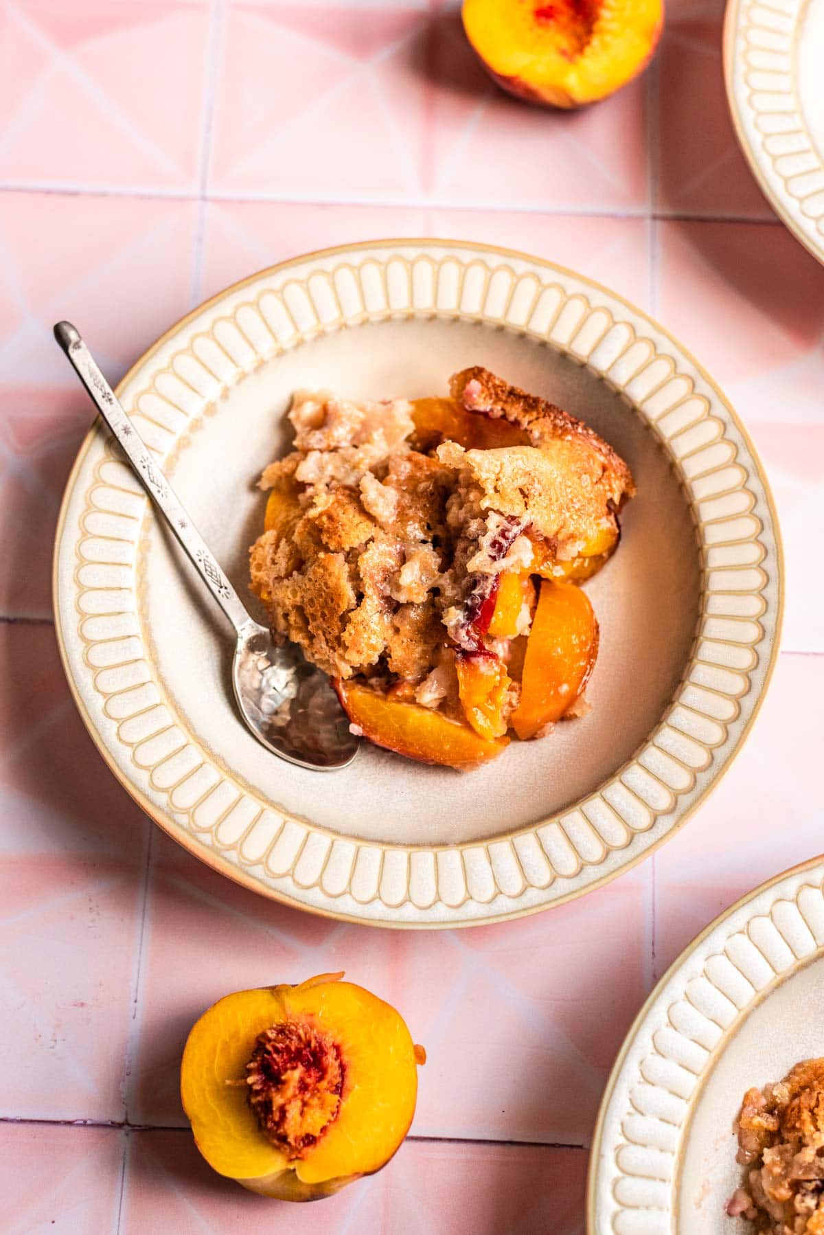 Cakey peach cobbler in a beige bowl on a pink tile table.