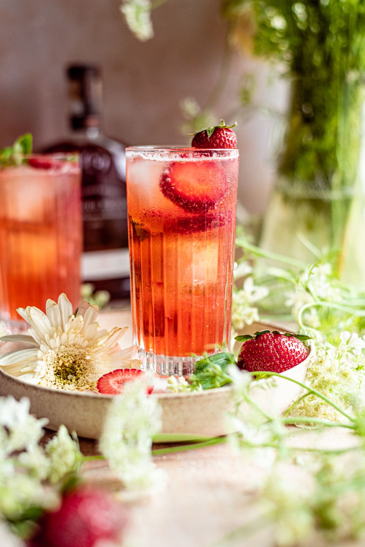 Strawberry bourbon cocktail in a highball glass garnished with strawberry slices and surrounded by white flowers.