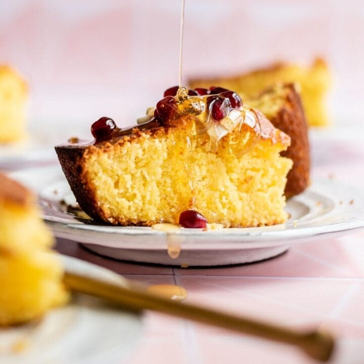 A slice of cornbread on a white plate with butter, honey, and pomegranate seeds.