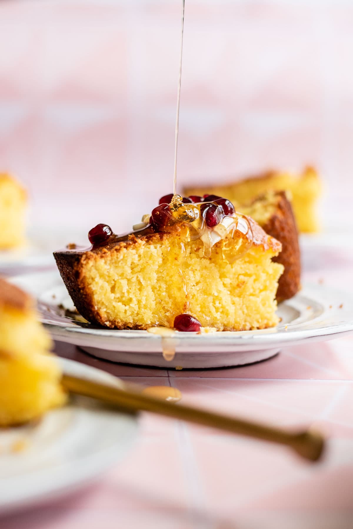 A slice of cornbread on a white plate with butter, honey, and pomegranate seeds.