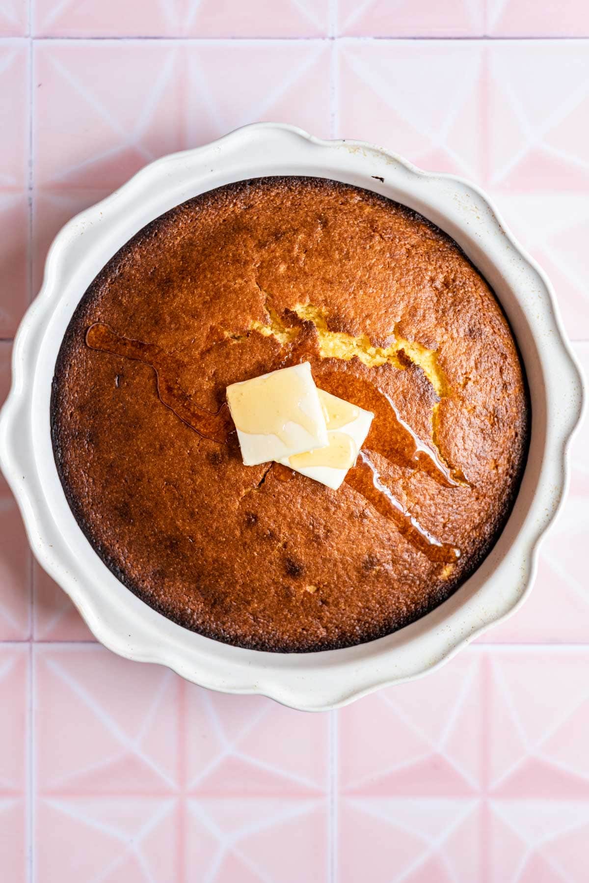 Jiffy cornbread in a circular baking dish with two slices of butter and a drizzle of honey on a pink tile table.
