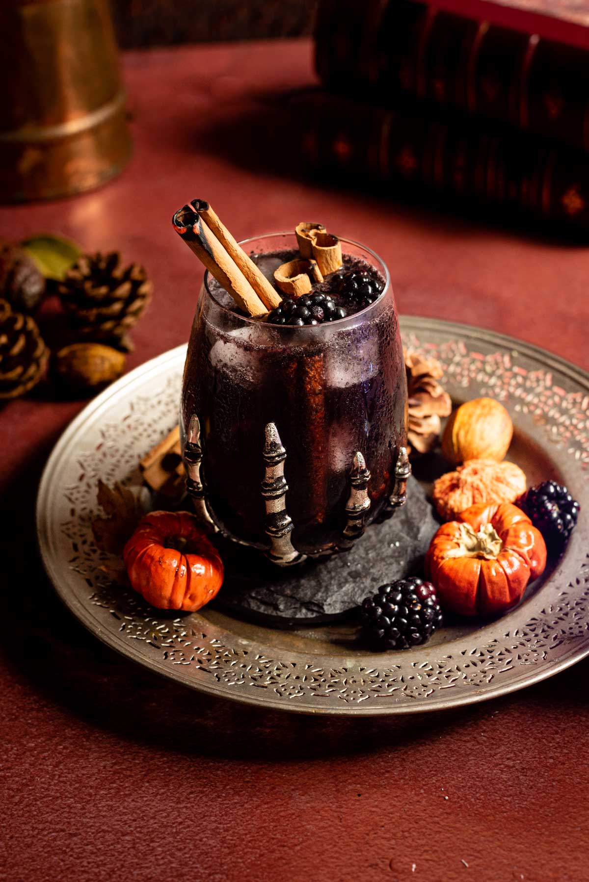The Black Flame Candle in a glass with a skeleton hand on a sliver plate with blackberries and small pumpkins.