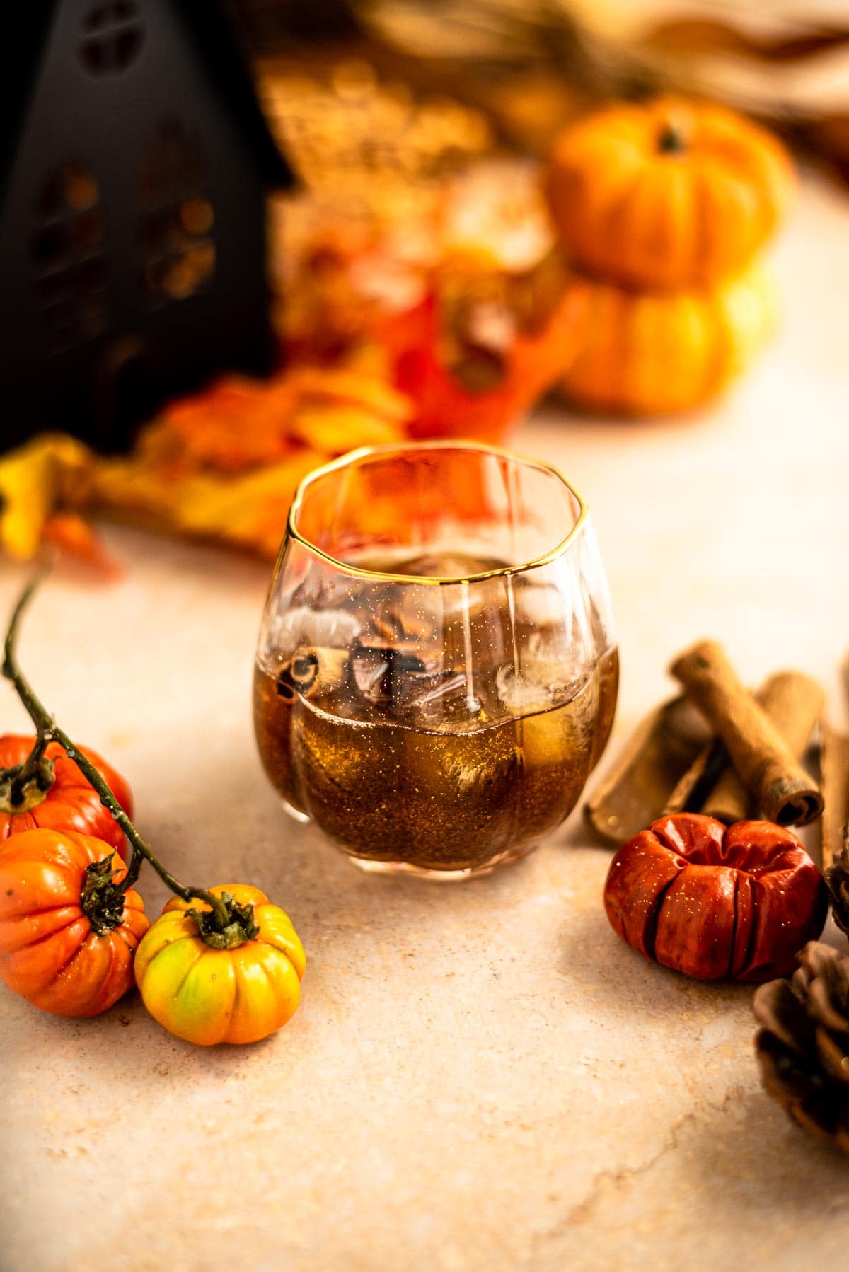 One cocktail with glitter, cinnamon sticks, and pecans in a pumpkin glass surrounded by mini pumpkins and leaves.