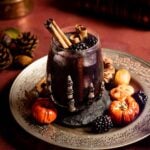The Black Flame Candle in a glass with a skeleton hand on a sliver plate with blackberries and small pumpkins.