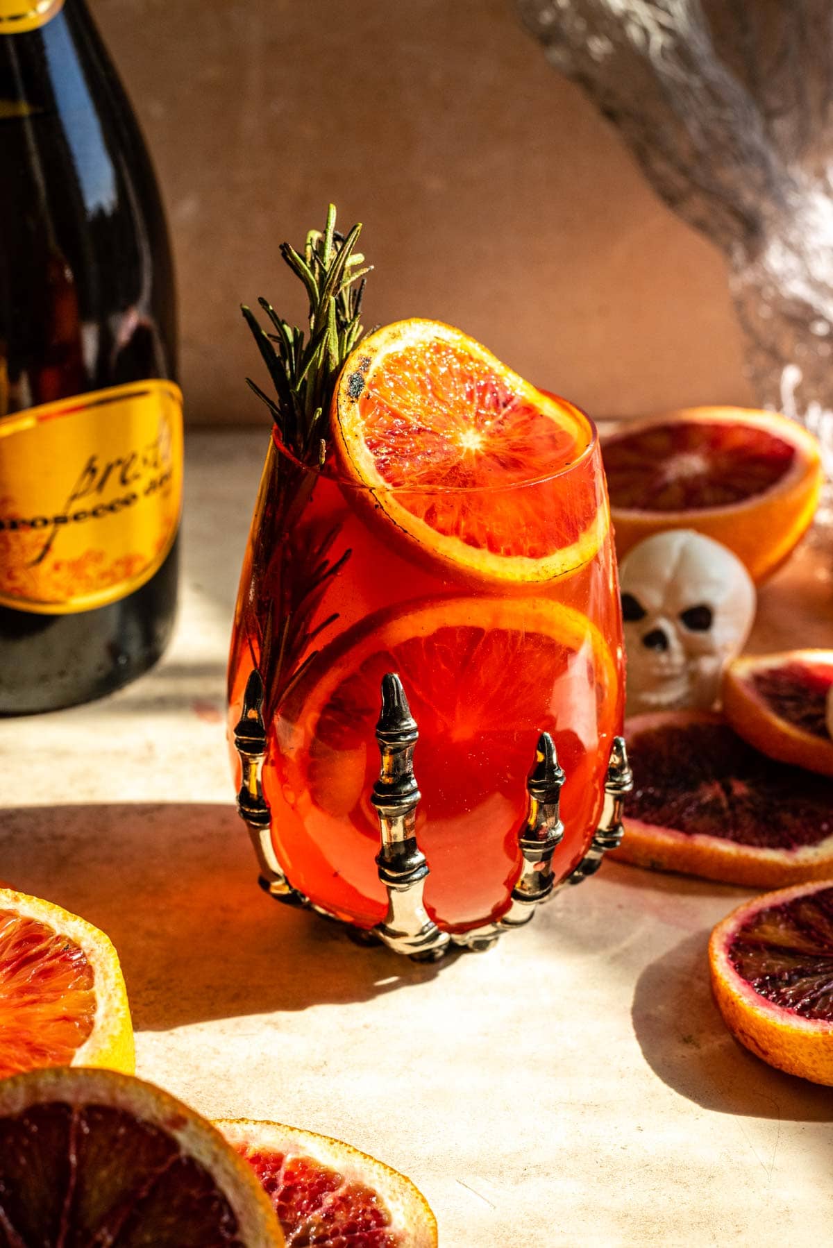 Blood orange spritz in a wine glass with a skeleton hand at the bottom. The cocktail is garnished with caramelized blood oranges an smoked rosemary. 