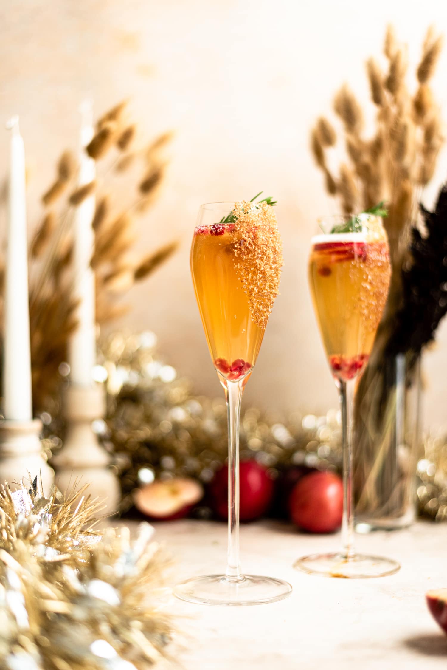 Two tulip champagne flutes rimmed with brown sugar and filled with apple cider mimosa. The cocktail is garnished with pomegranate seeds, apple slices, and rosemary.  