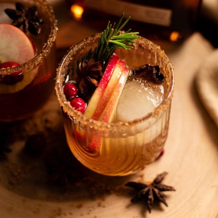 Overhead shot of an apple cider old fashioned garnished with 3 slices of apples, cranberries, cinnamon and rosemary.