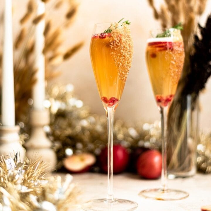 Two tulip champagne flutes rimmed with brown sugar and filled with apple cider mimosa. The cocktail is garnished with pomegranate seeds, apple slices, and rosemary.