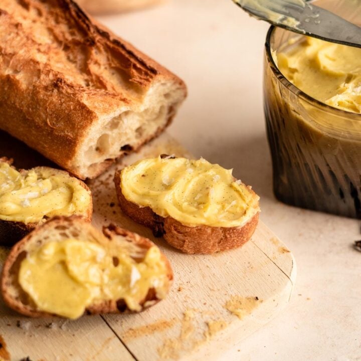 A glass filled with vanilla butter, with a sliced baguette on a cutting board with butter smeared on the slices.