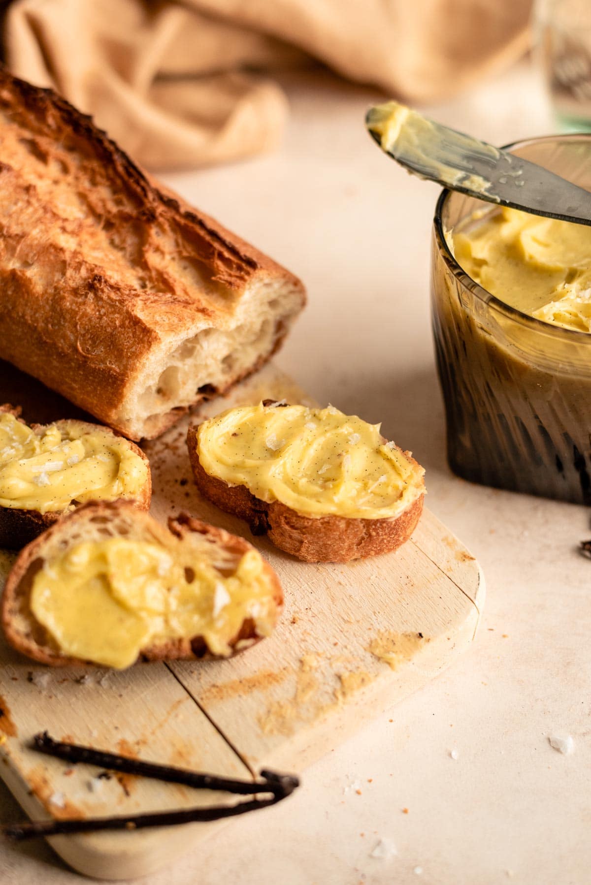 A glass filled with vanilla butter, with a sliced baguette on a cutting board with butter smeared on the slices.