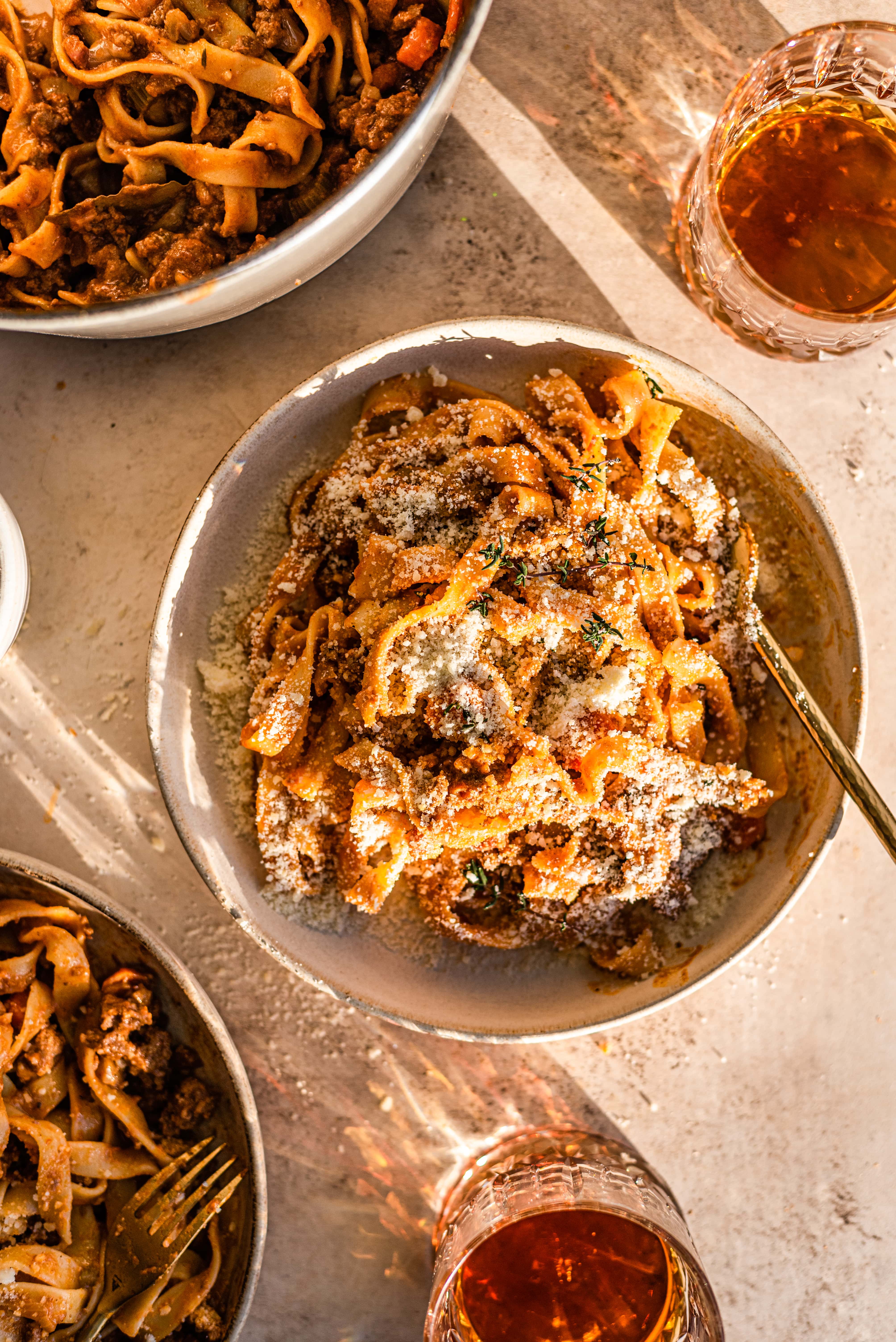 A brown bowl full of bolognese pasta, sprinkled with parma cheese, thyme, and with two glasses of bourbon above and below.