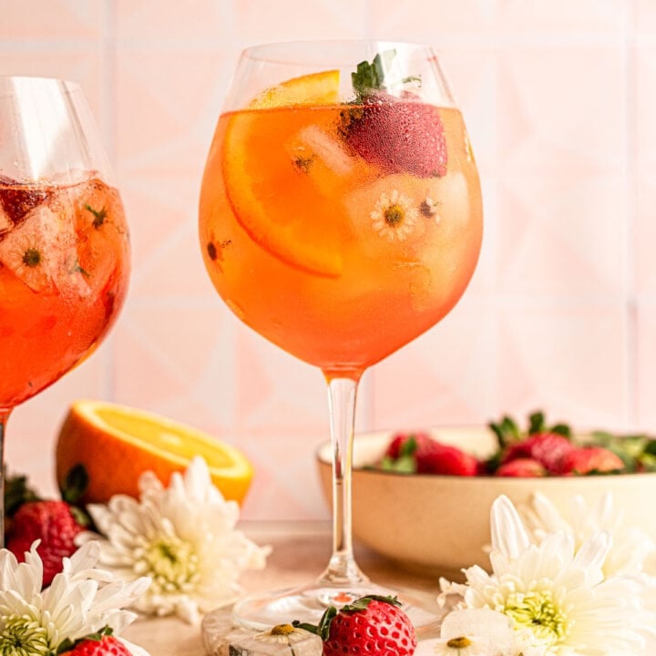 A strawberry Aperol Spritz on a brown table and garnished with flower ice, orange slice, and a strawberry.