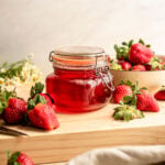 Strawberry simple syrup in an airtight jar and sitting on a wood cutting board. Strawberries are sitting all around the syrup.