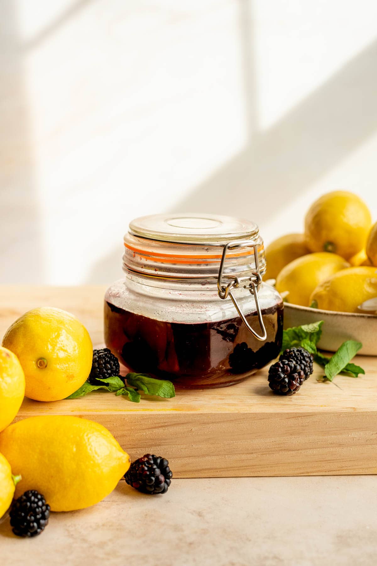 Blackberry simple syrup in a glass container, sitting on a large wood cutting board surrounded by blackberries and lemons.