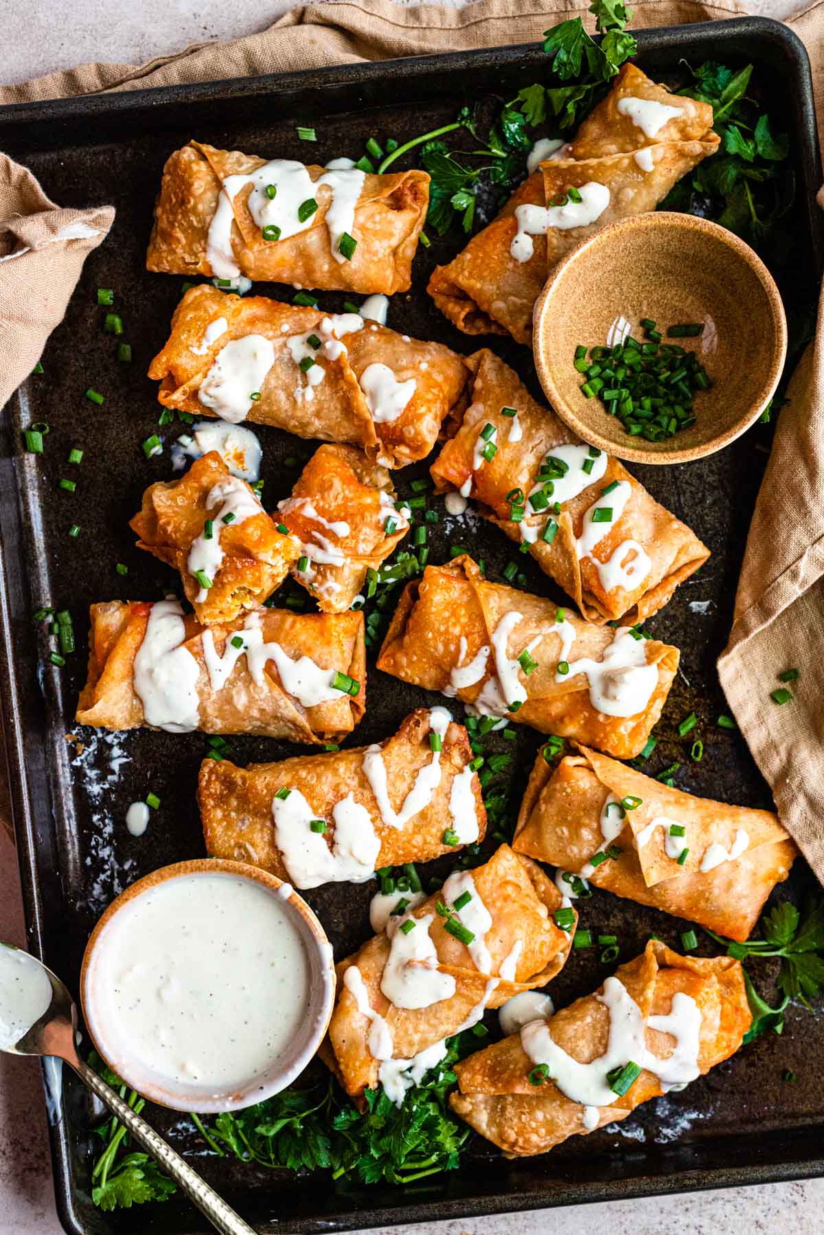 14 buffalo chicken egg rolls on a baking sheet with two little bowls one with blue cheese dressing and one full of chives.