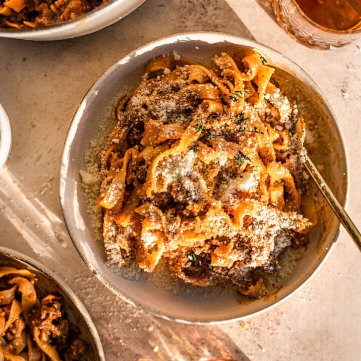 A brown bowl full of bolognese pasta, sprinkled with parma cheese, thyme, and with two glasses of bourbon above and below.
