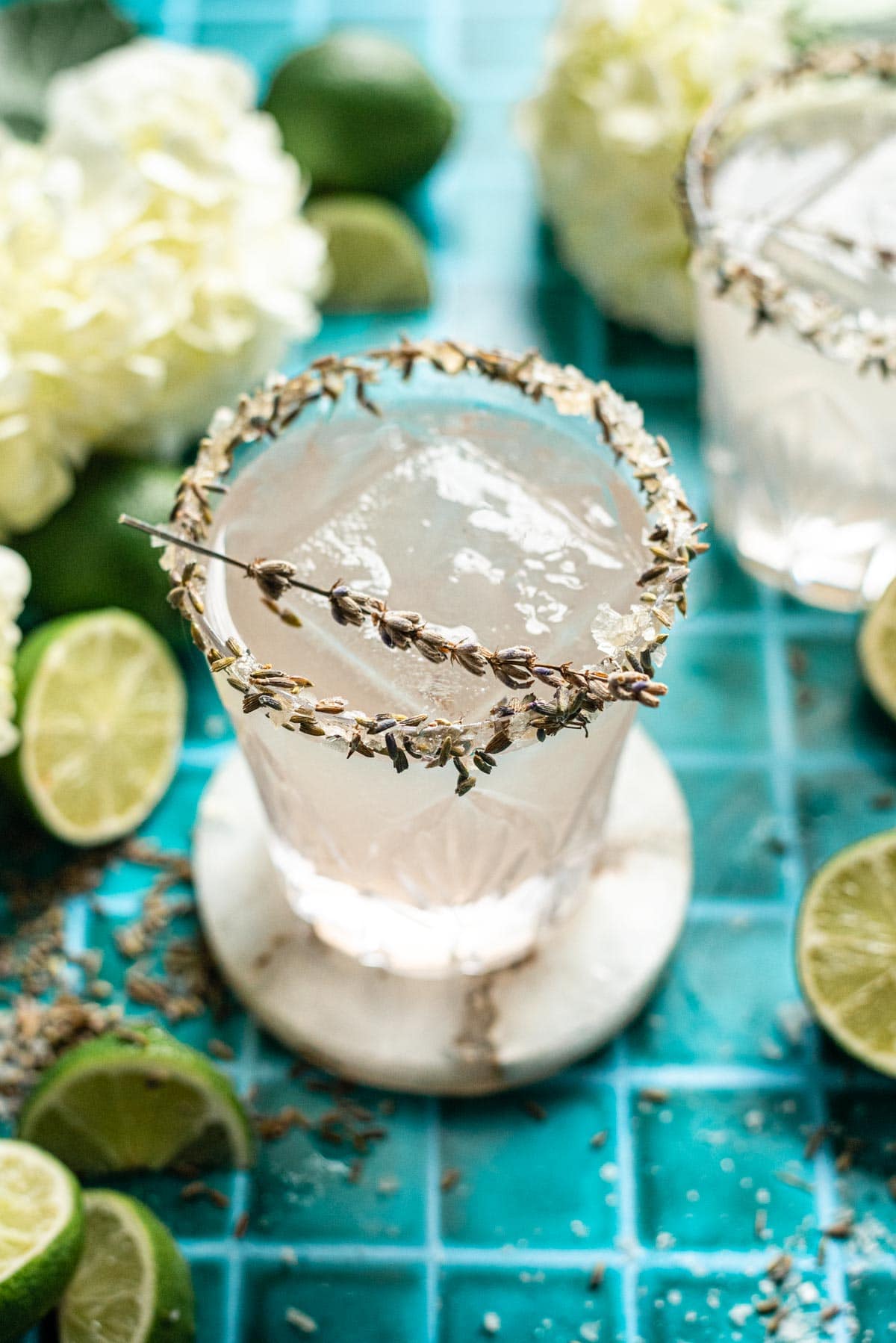 Lavender margarita in a cut glass with a salt and lavender rim on a teal tile table.