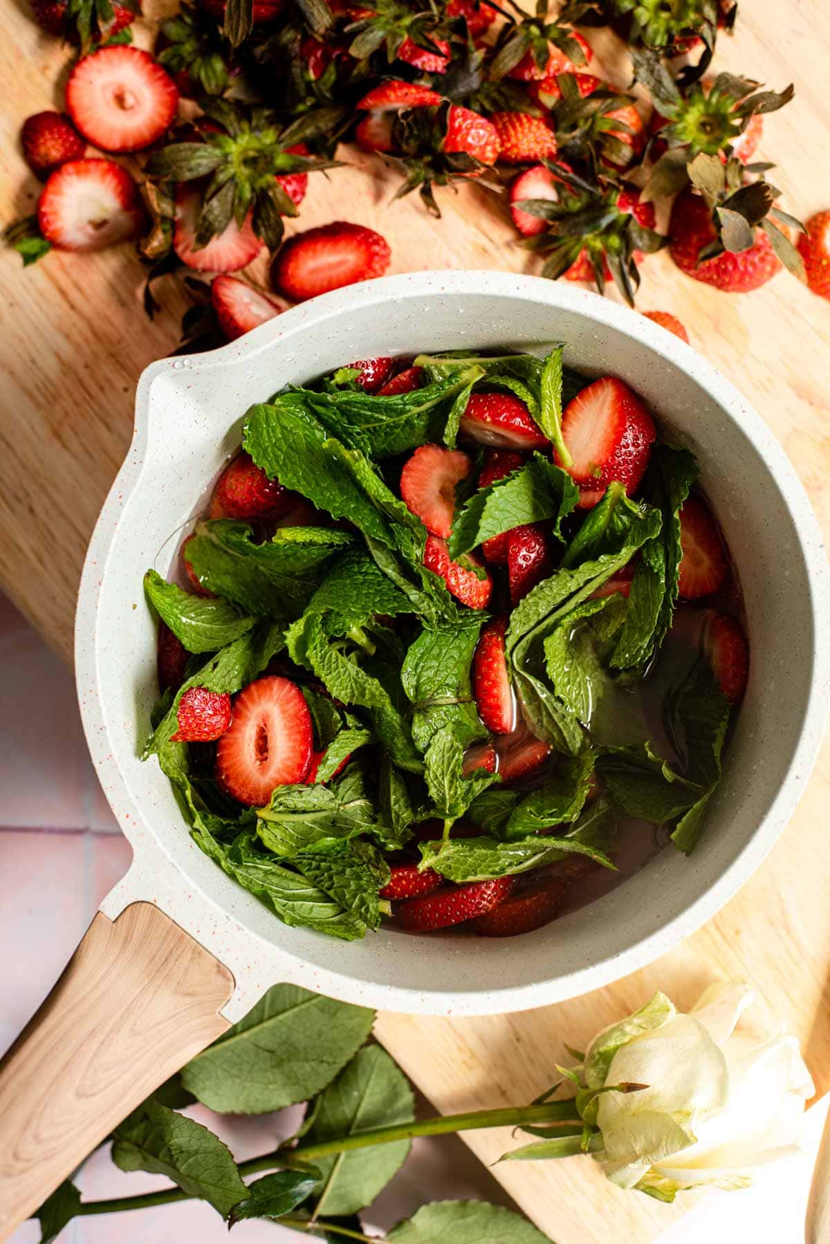 Fresh mint, cut strawberries, sugar, and water in a white saucepan.