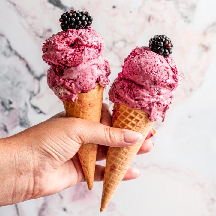 A hand holding two ice cream cones filled with blackberry ice cream with two blackberries on top.