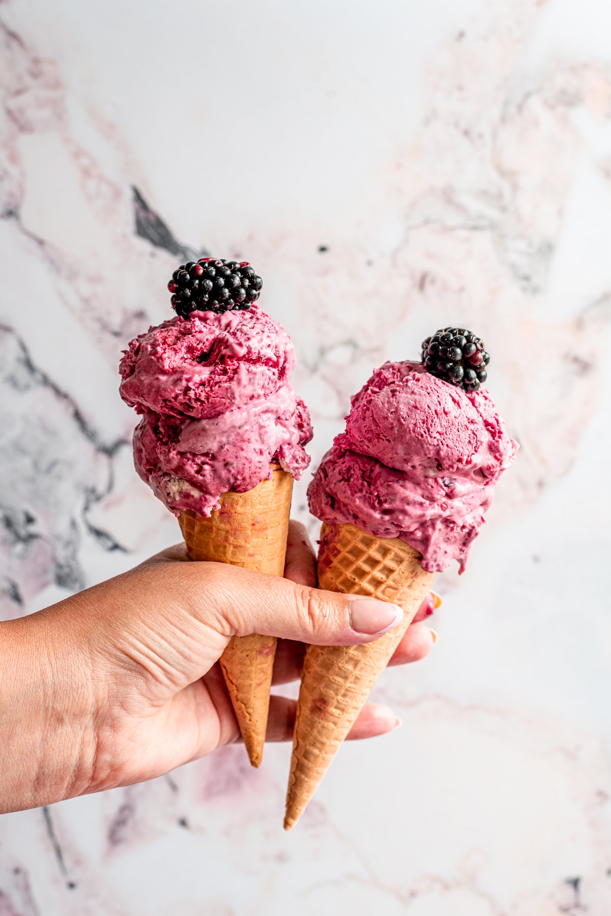 A hand holding two ice cream cones filled with blackberry ice cream with two blackberries on top.