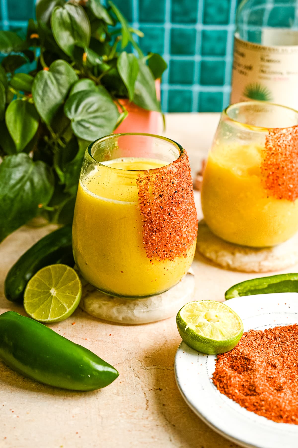A frozen spicy mango margarita in a glass rimmed with tajin on a brown marble table and green tile background.