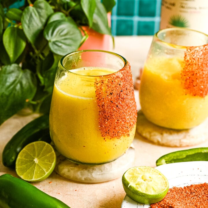 A frozen spicy mango margarita in a glass rimmed with tajin on a brown marble table and green tile background.