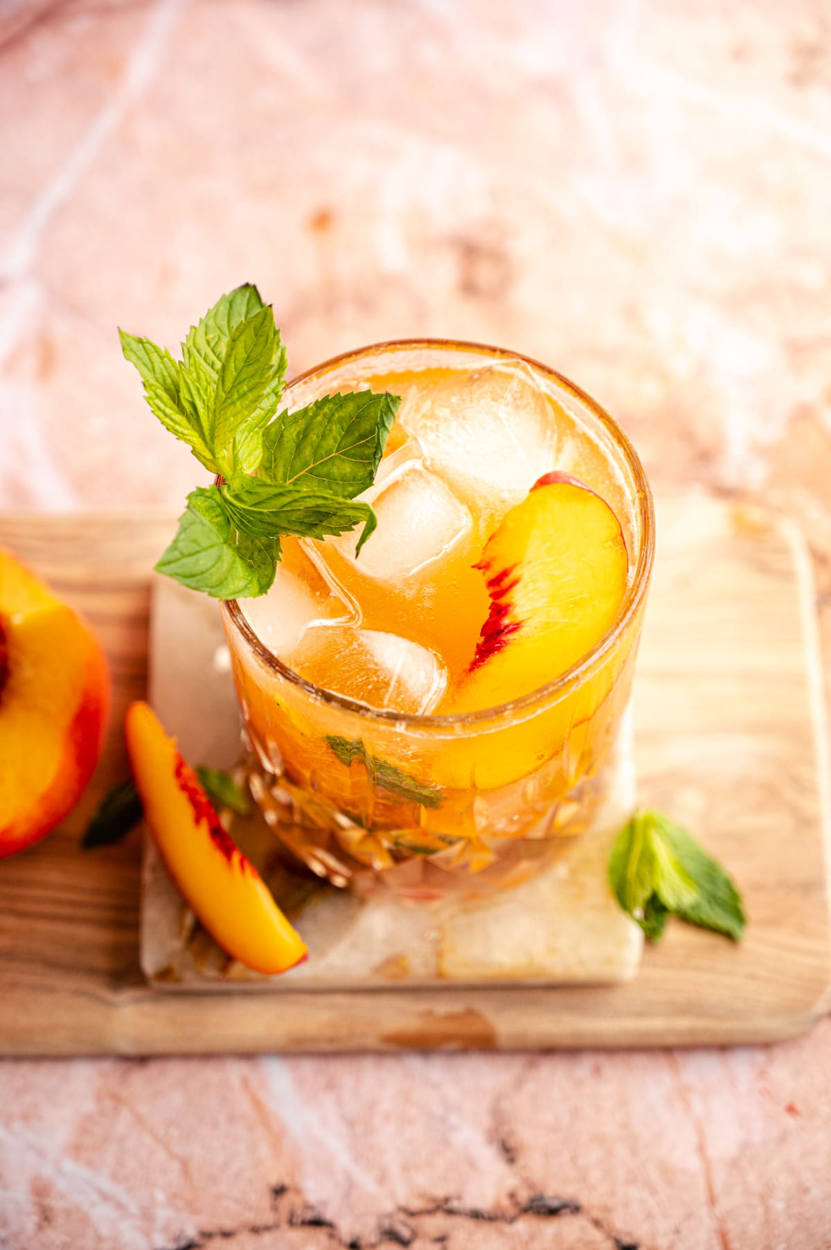 Over head shot of an old fashioned glass filled with a peach smash and garnished with peach slices and a mint sprig. Its on a marble coaster and a pink marble table.