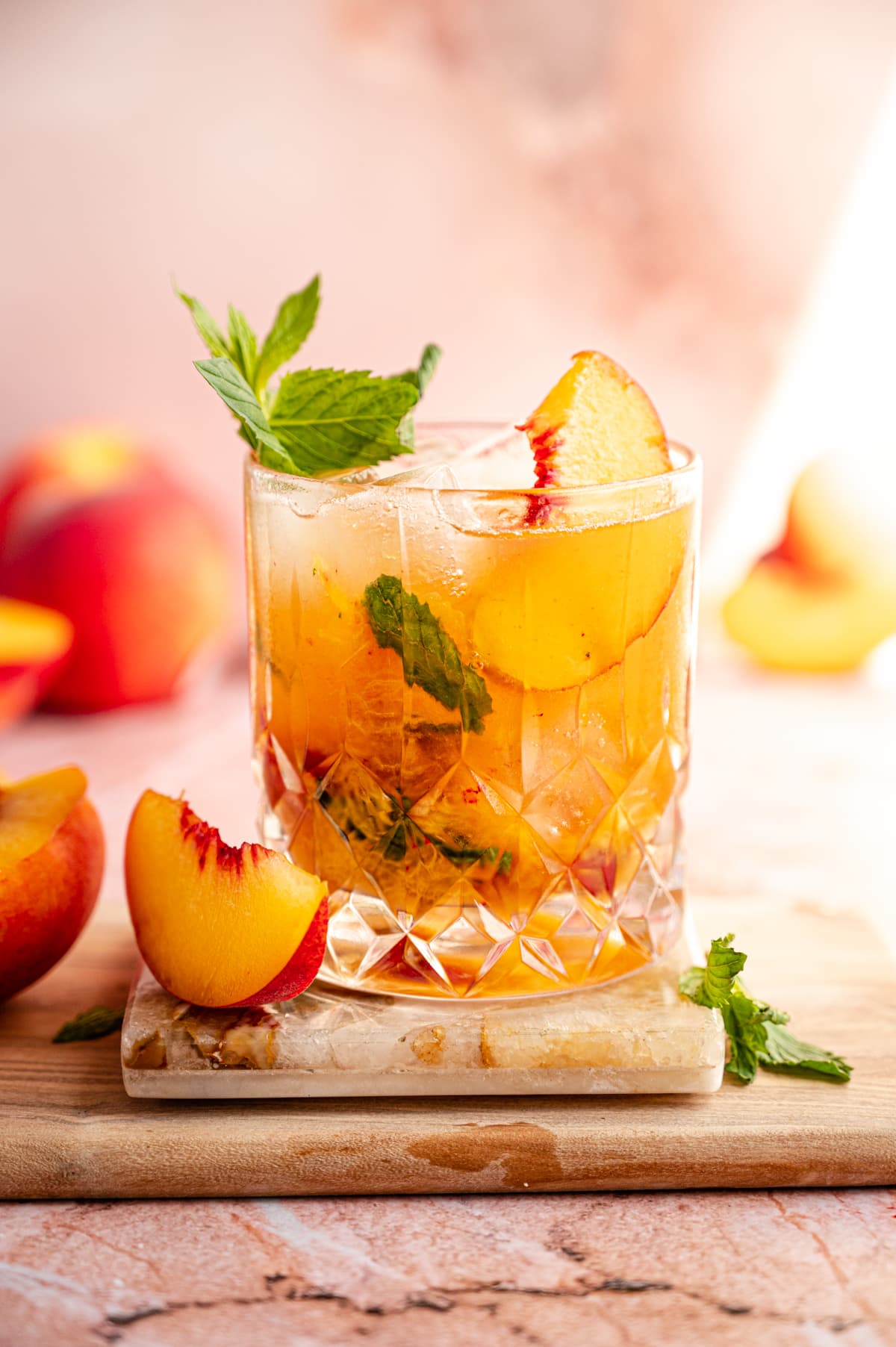 An old fashioned glass filled with a peach smash and garnished with peach slices and a mint sprig. Its on a marble coaster and a pink marble table.