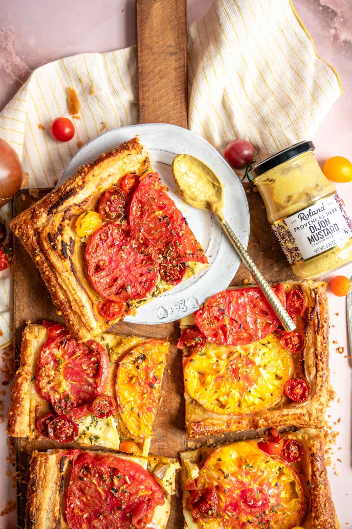 A sliced tomato tart on a wood cutting board with one grey dessert plate with a slice sitting on top. A jar of Roland Foods Mustard is off to the side.